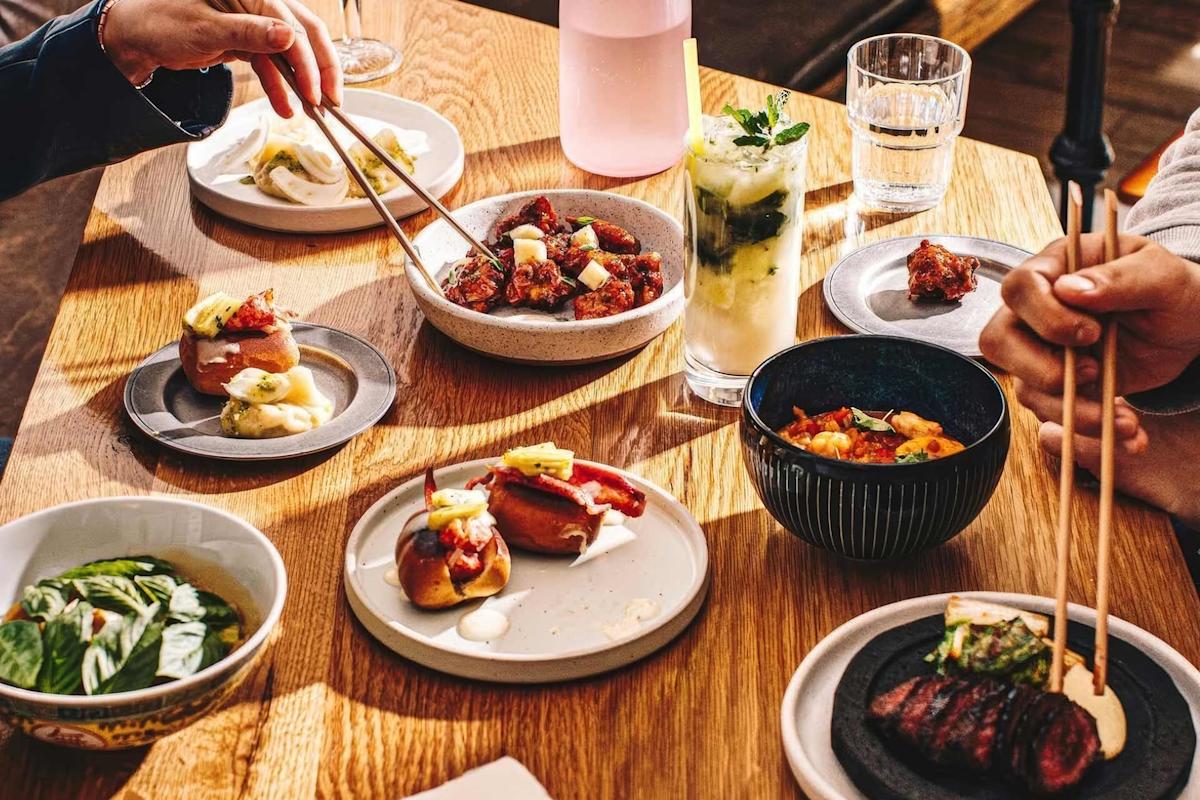 A wooden table set with assorted dishes, including sushi, small plates of appetizers, a bowl of greens, a cocktail, and water. Two people are using chopsticks to pick up food. Sunlight streams across the scene.