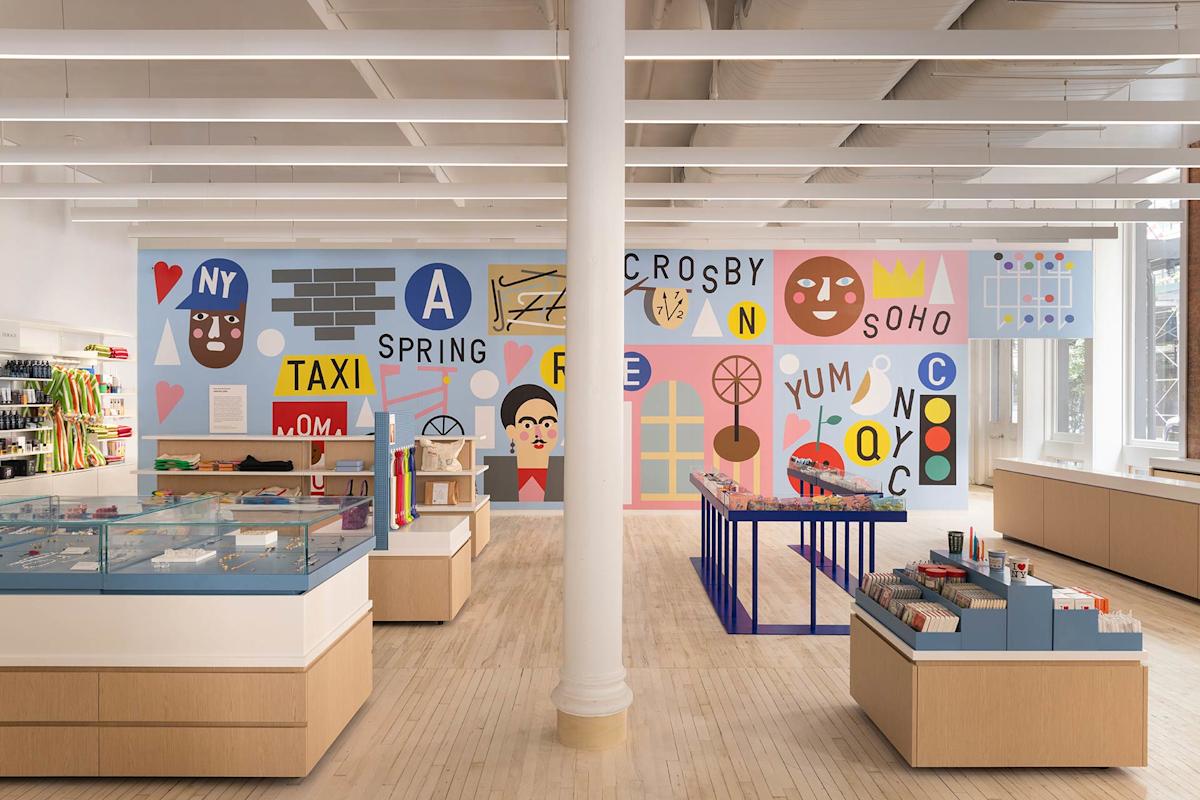 A bright, modern store with wooden floors, displays of books and gifts, and a colorful mural featuring abstract faces, letters, and New York-themed illustrations on the back wall.