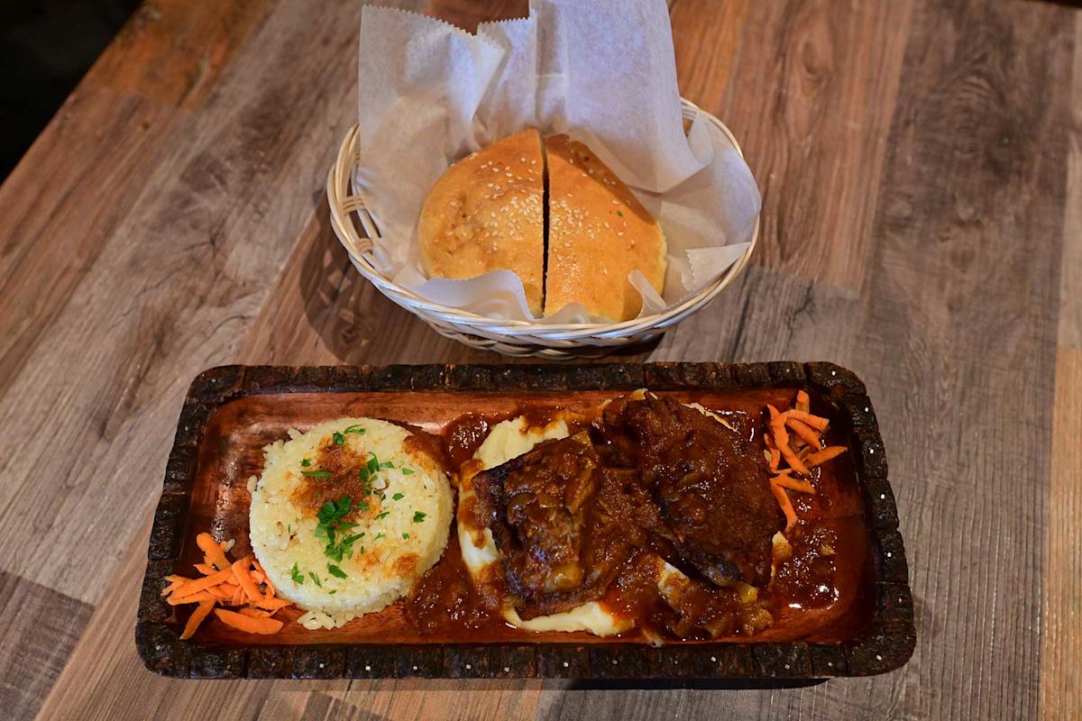 A wooden platter with rice, mashed potatoes, beef stew, and shredded carrots, served alongside a basket with two pieces of sesame-seeded bread on a rustic wooden table.