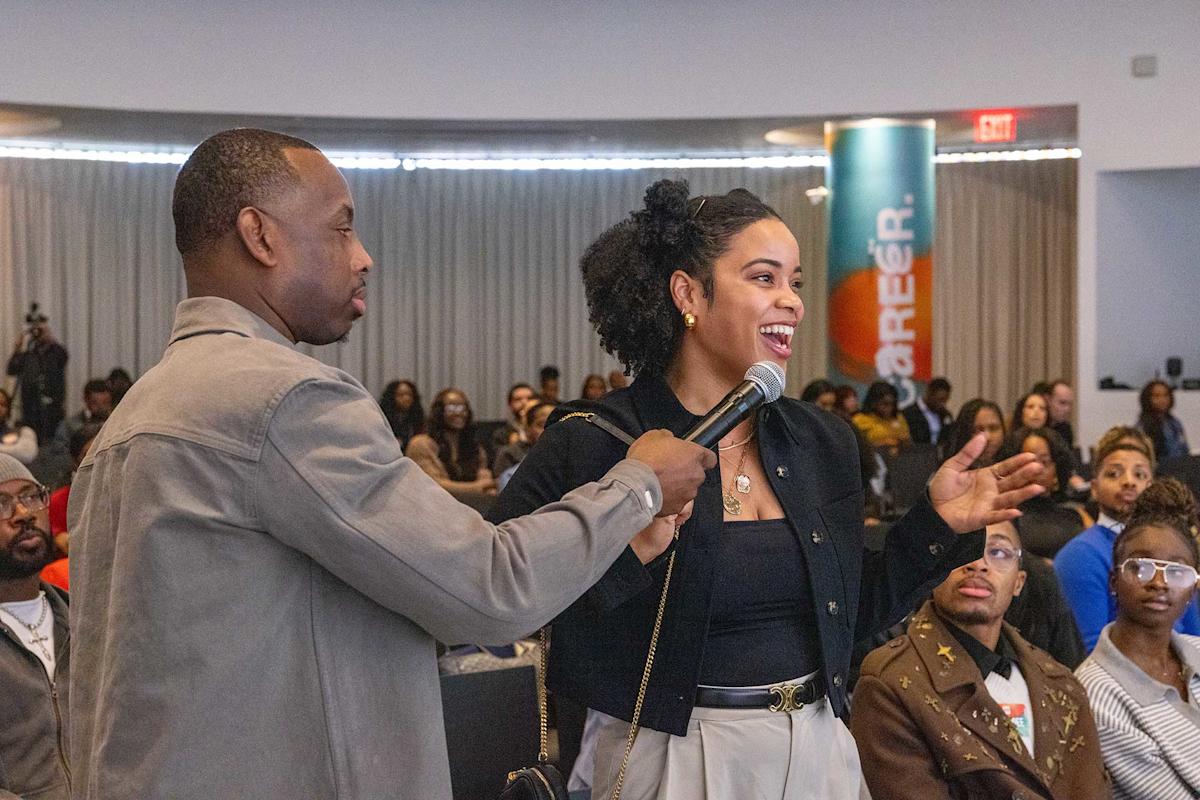 A man holds a microphone for a smiling woman who is speaking to an audience in a large room. People sit and watch in the background, and a colorful banner is visible on the right side.
