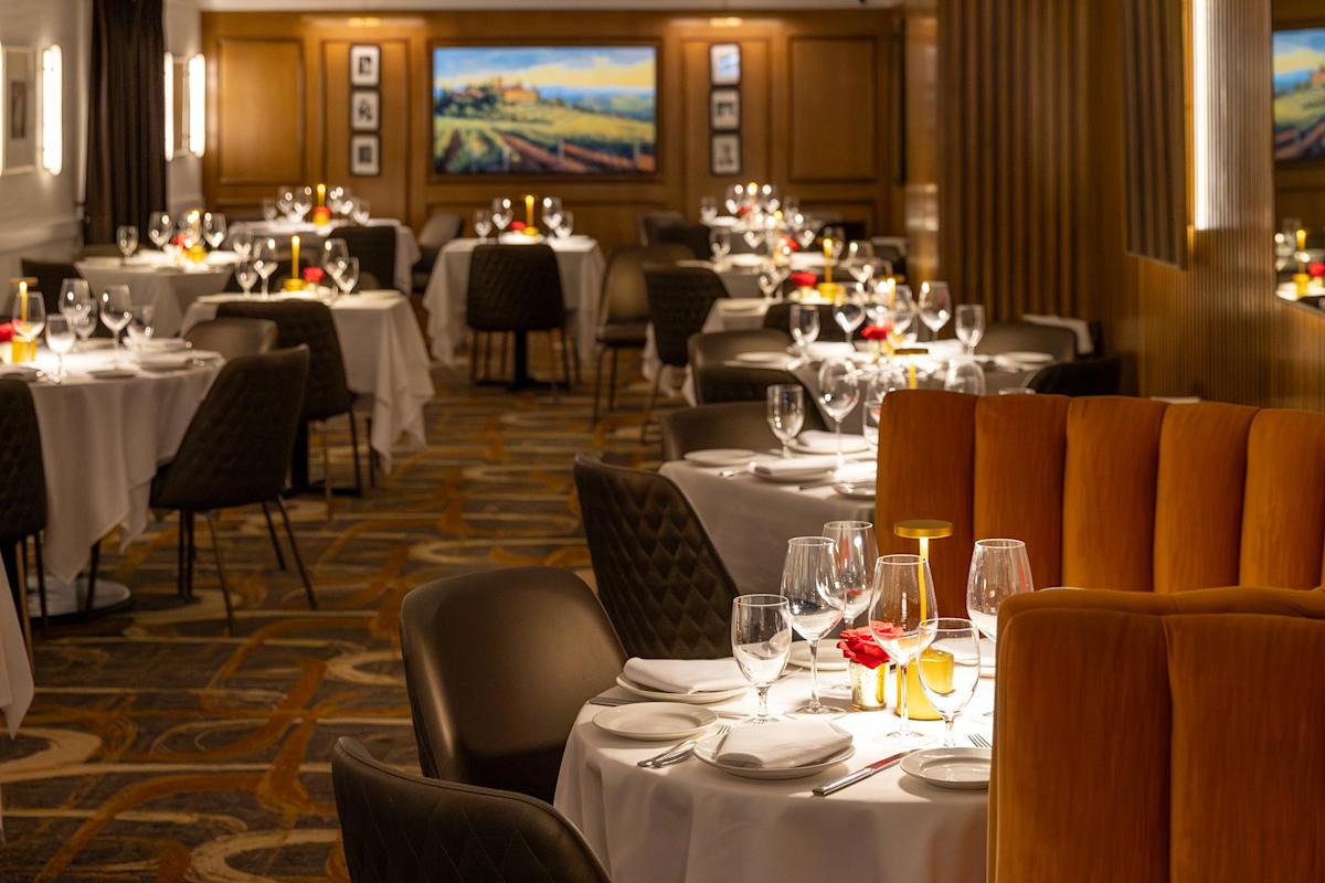 Elegant restaurant interior with white tablecloths, set tables with wine glasses, candles, and red flower centerpieces. Warm lighting and paintings on wood-paneled walls create a cozy, upscale ambiance.