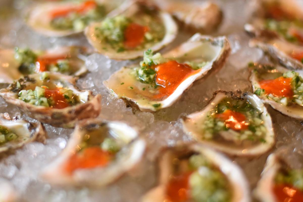 Fresh oysters served on a bed of crushed ice, each topped with a green herb mixture and a dollop of bright orange sauce.