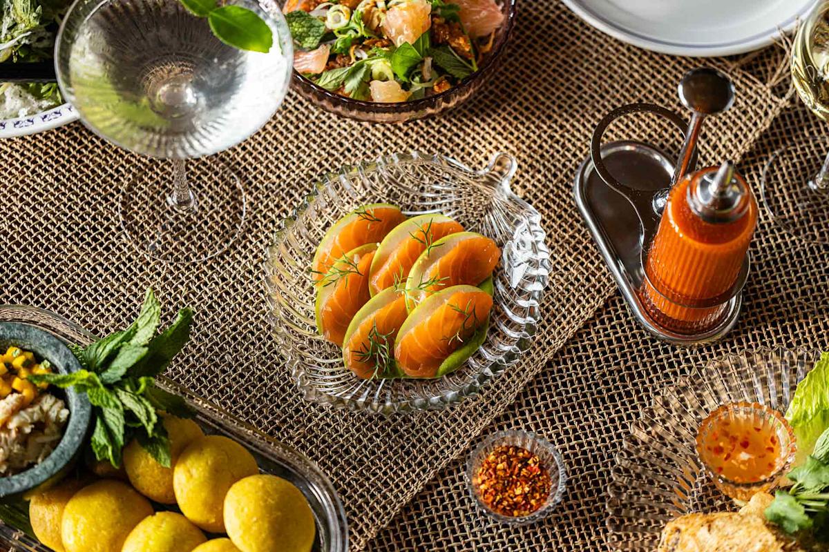 A glass dish with four salmon sashimi pieces garnished with herbs is on a woven tablecloth, surrounded by various dishes, a drink, condiments, and fresh greens.