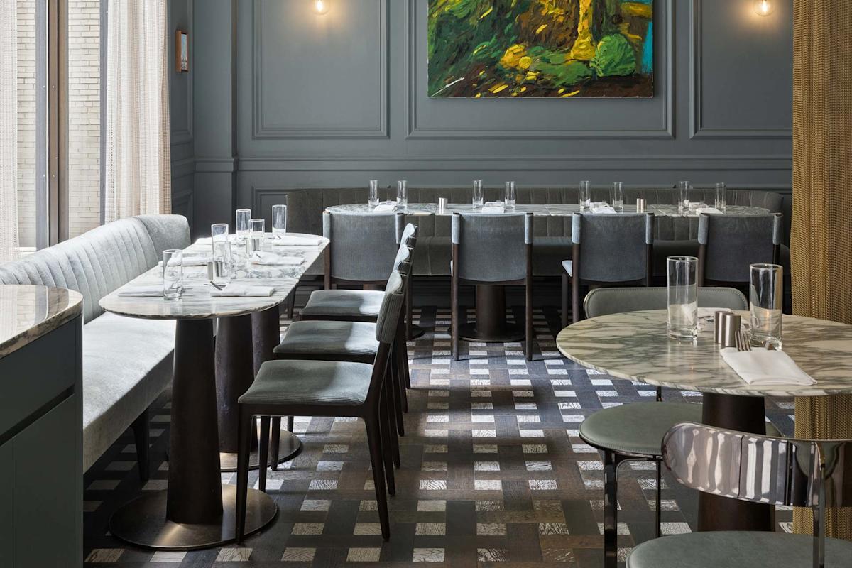A modern restaurant interior with gray walls, a marble-top table, gray upholstered chairs, a long cushioned bench, glassware on tables, and a large abstract painting on the wall. Geometric patterned flooring is visible.