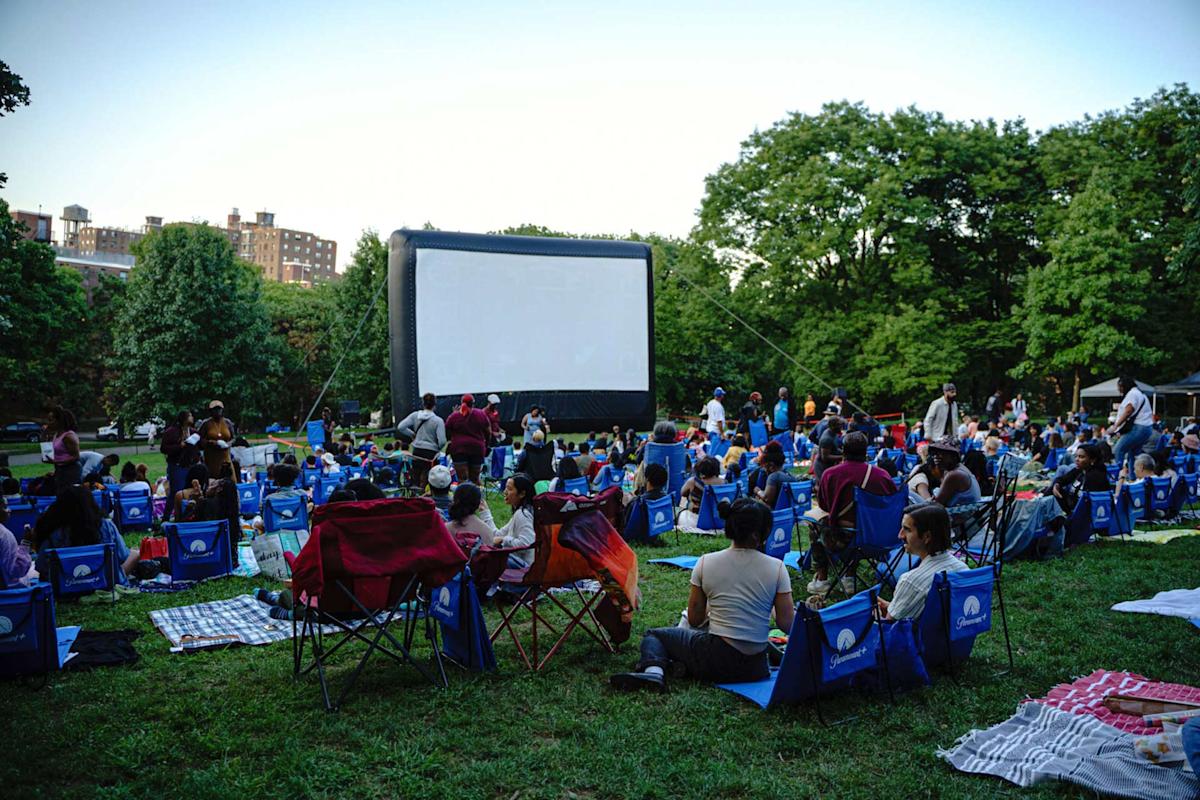 A crowd of people sits on lawn chairs and blankets in a grassy park, facing a large outdoor movie screen, surrounded by trees under a clear sky.