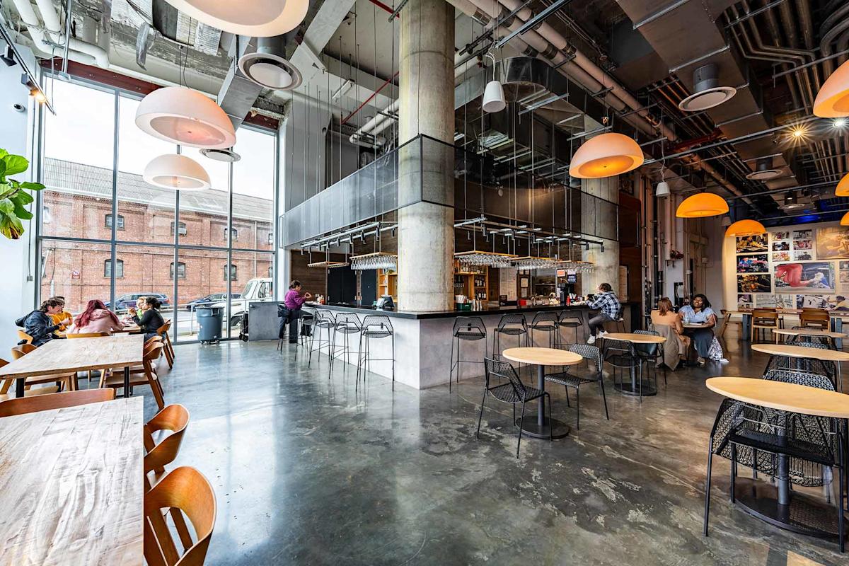A modern cafe with high ceilings, large windows, hanging lights, and industrial decor. People sit at wooden and metal tables, chatting and working. The bar area is central, with exposed pipes overhead.