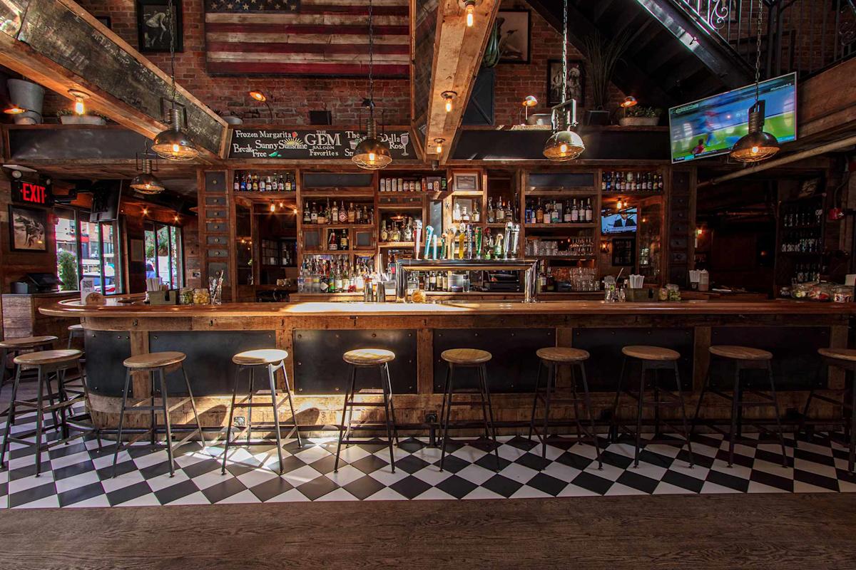 A rustic bar with wooden stools, a polished wooden counter, shelves stocked with bottles, hanging pendant lights, a checkered floor, and an American flag on the brick wall above the bar. A TV is on in the upper right corner.