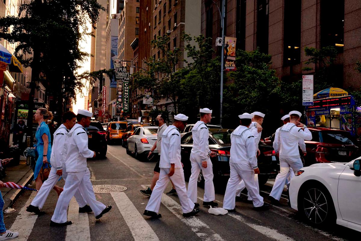 A group of sailors in white uniforms cross a busy street in an urban setting, with cars and tall buildings on either side. The scene captures lively city life and the sun setting in the background.