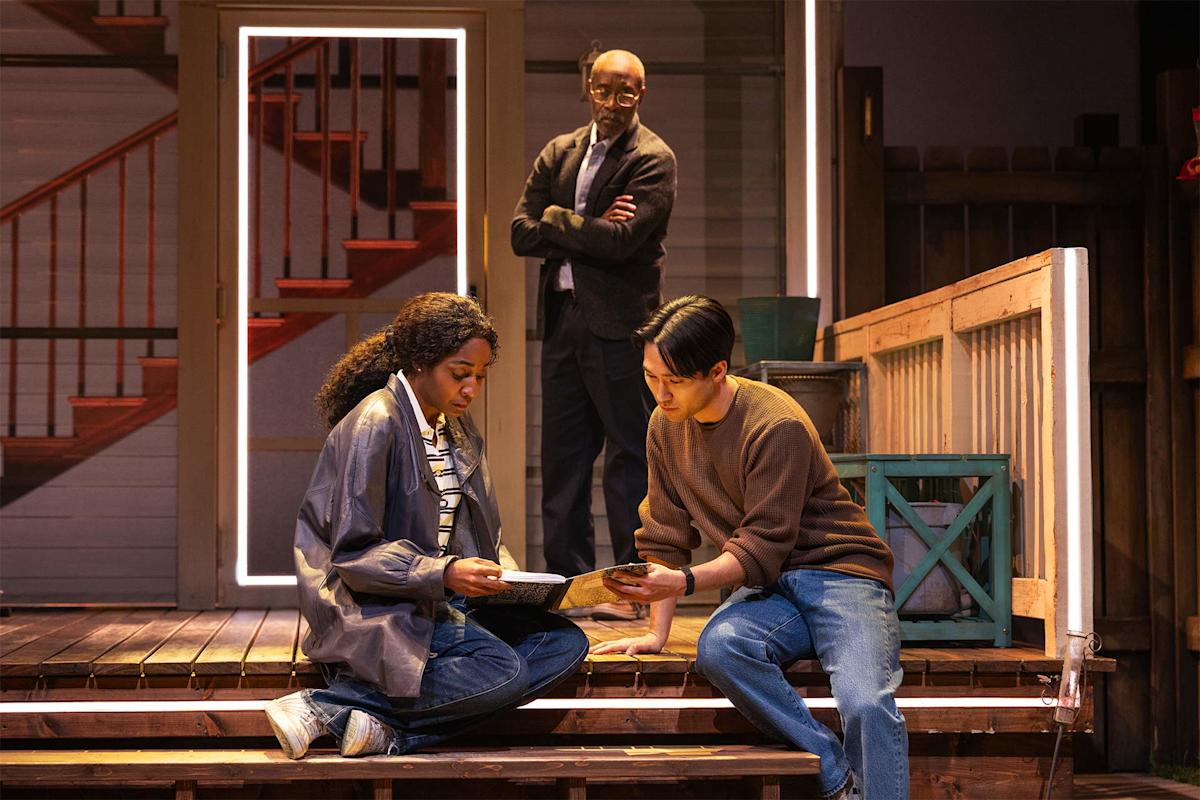 Two people sit on porch steps examining a document, while a man stands behind them with arms crossed, watching. The scene appears to be from a stage play set in front of a house with stairs in the background.