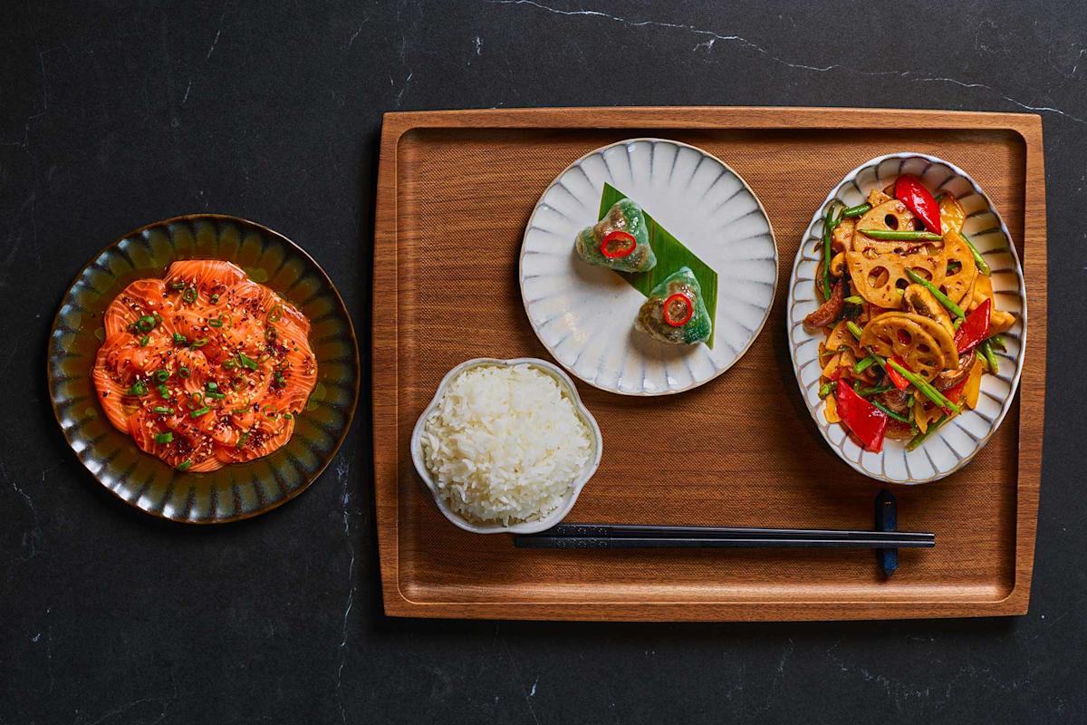 A wooden tray holds a bowl of white rice with chopsticks, a plate of sliced lotus root with peppers, a small dish with a wrapped bite, and a separate plate of sliced salmon topped with herbs. All dishes are neatly arranged.