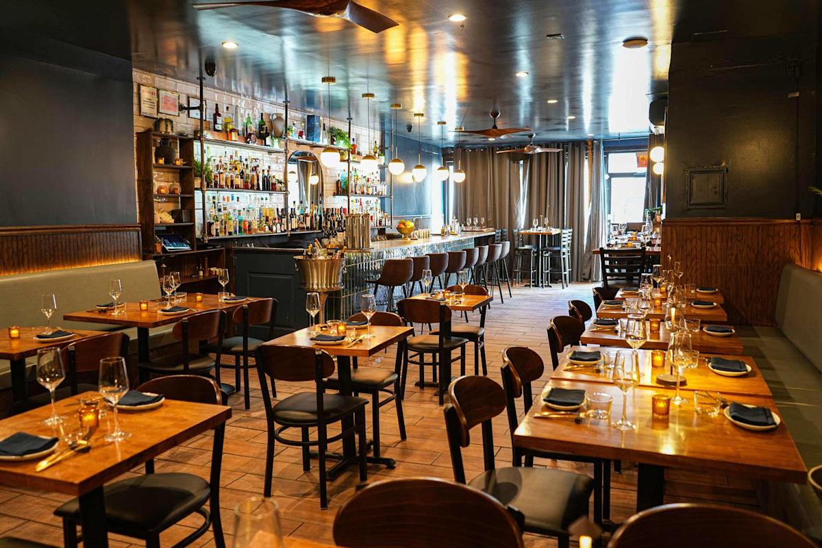 A stylish, modern restaurant interior with wooden tables and chairs set for dining, a long bar with stools, shelves of bottles, and warm lighting creating an inviting atmosphere.