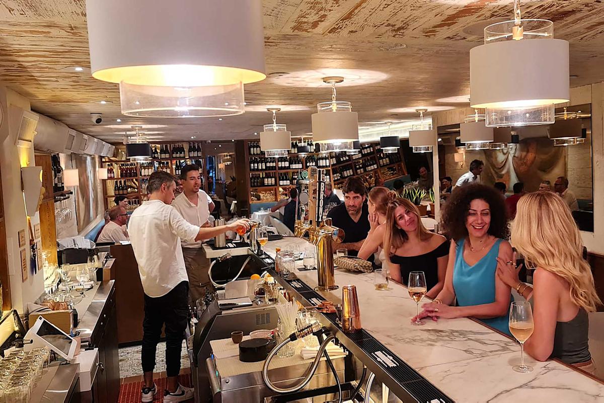 A lively bar scene with people sitting and chatting at a marble counter, bartenders preparing drinks, and shelves of wine bottles lining the walls in a warmly lit, modern interior.