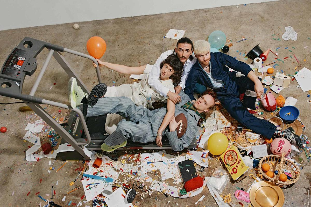 Four people in colorful clothes lounge on and around a treadmill surrounded by scattered party items, balloons, confetti, snacks, and various playful clutter on a concrete floor.