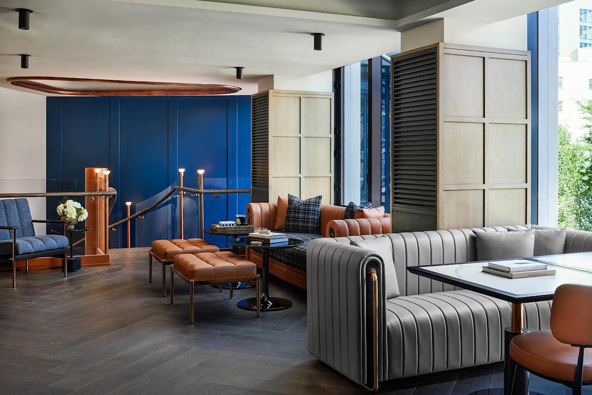 Modern lounge area with stylish furniture, including a gray sofa, brown leather benches, armchairs, and a white table. Blue accent wall, large windows, and wooden flooring add to the sophisticated, contemporary feel.
