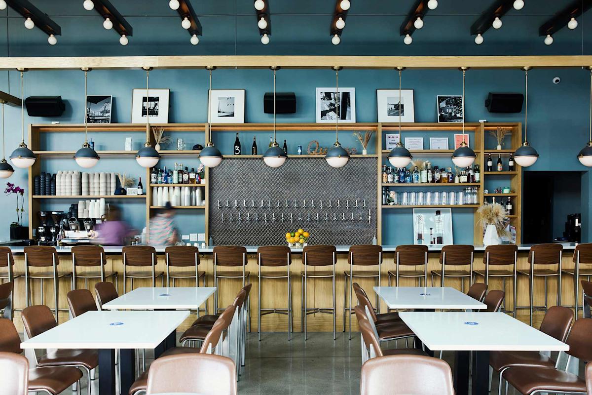 Modern bar with a long wooden counter, brown chairs, and white tables. Pendant lights hang from the ceiling. Shelves behind the bar display bottles, framed photos, and decor. Two people are blurred, moving behind the counter.