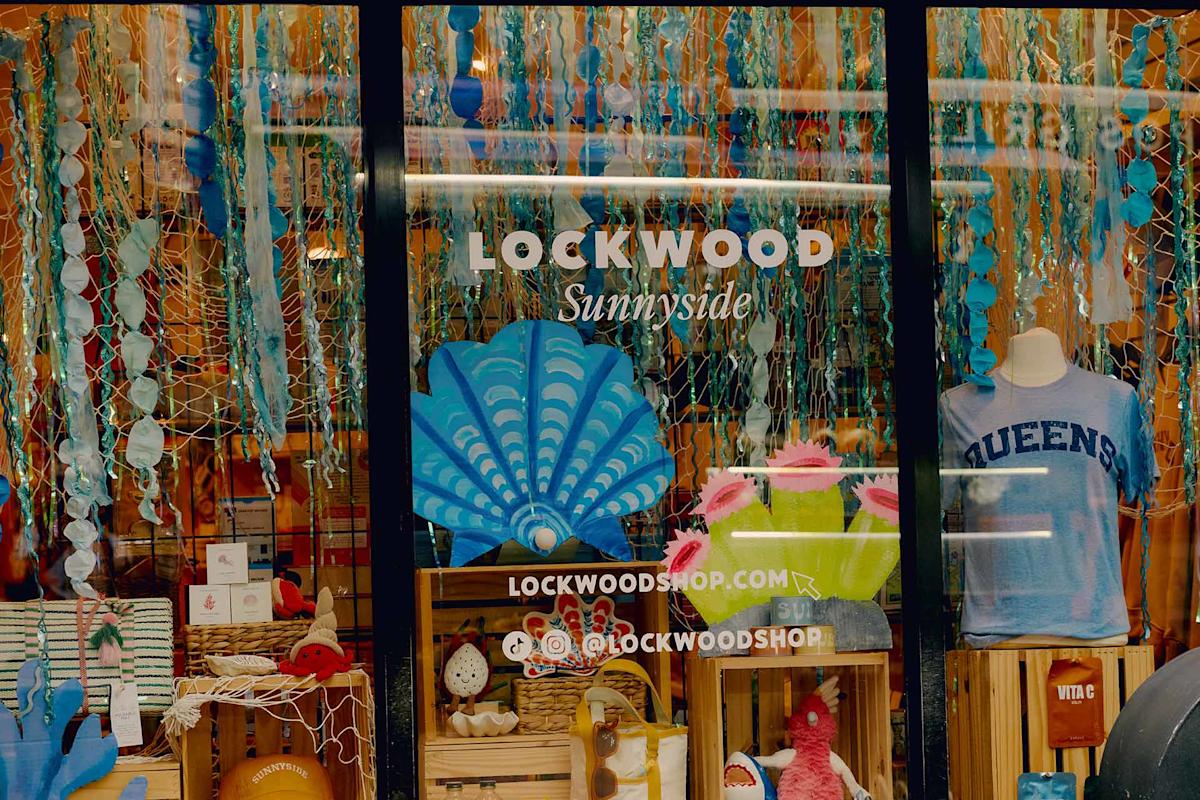 A colorful shop window display at Lockwood Sunnyside features hanging blue and turquoise decorations, a large shell cutout, gift items, books, and a mannequin wearing a light blue "QUEENS" t-shirt.