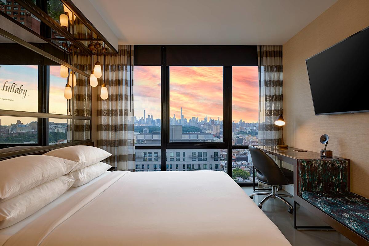 A modern hotel room with a large bed, sleek desk, and wall-mounted TV; floor-to-ceiling windows reveal a city skyline and colorful sunset sky outside.