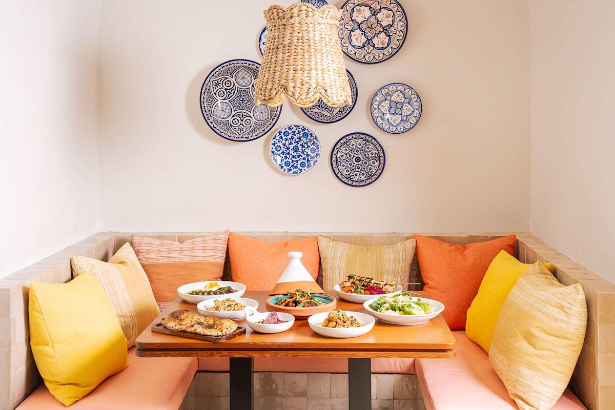 A cozy dining nook with colorful cushions and a wooden table set with various Moroccan dishes. Blue patterned plates decorate the white wall above, and a woven pendant lamp hangs overhead.