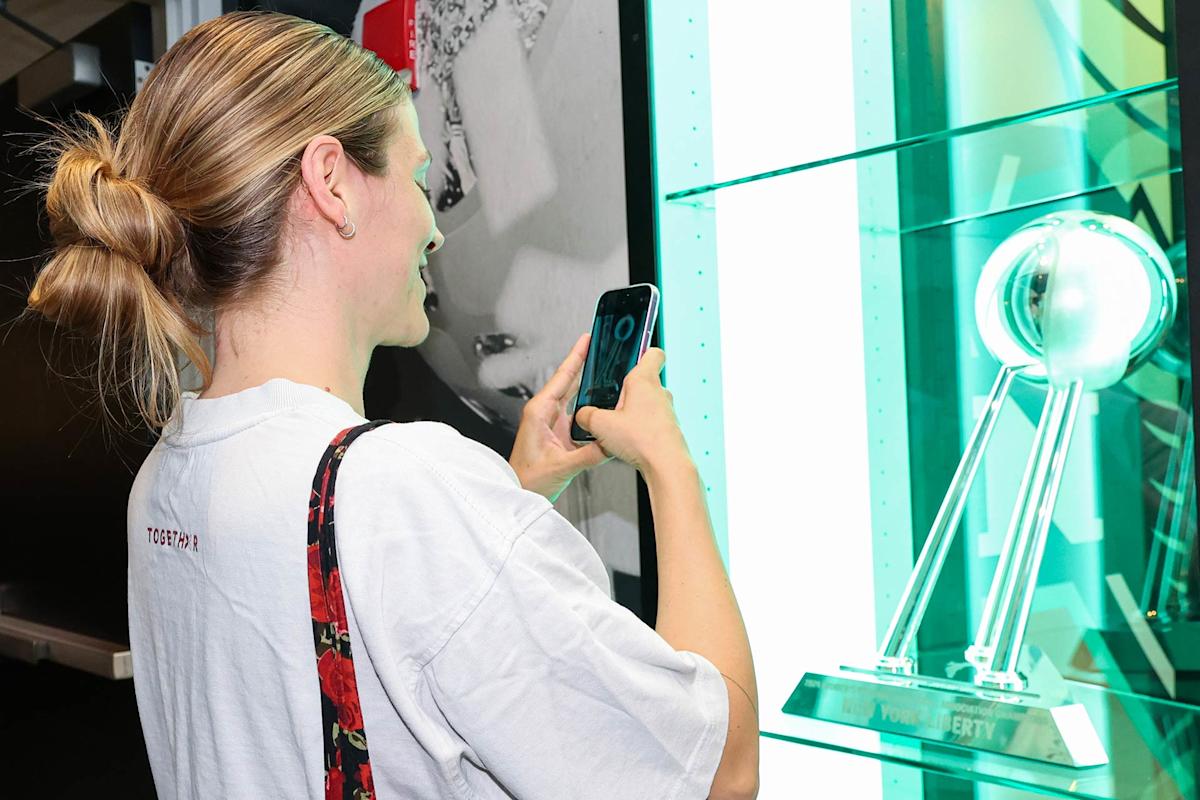 A woman with blonde hair in a bun takes a photo of a shiny silver trophy displayed in a glass case. She is wearing a white T-shirt and carrying a floral bag over her shoulder.