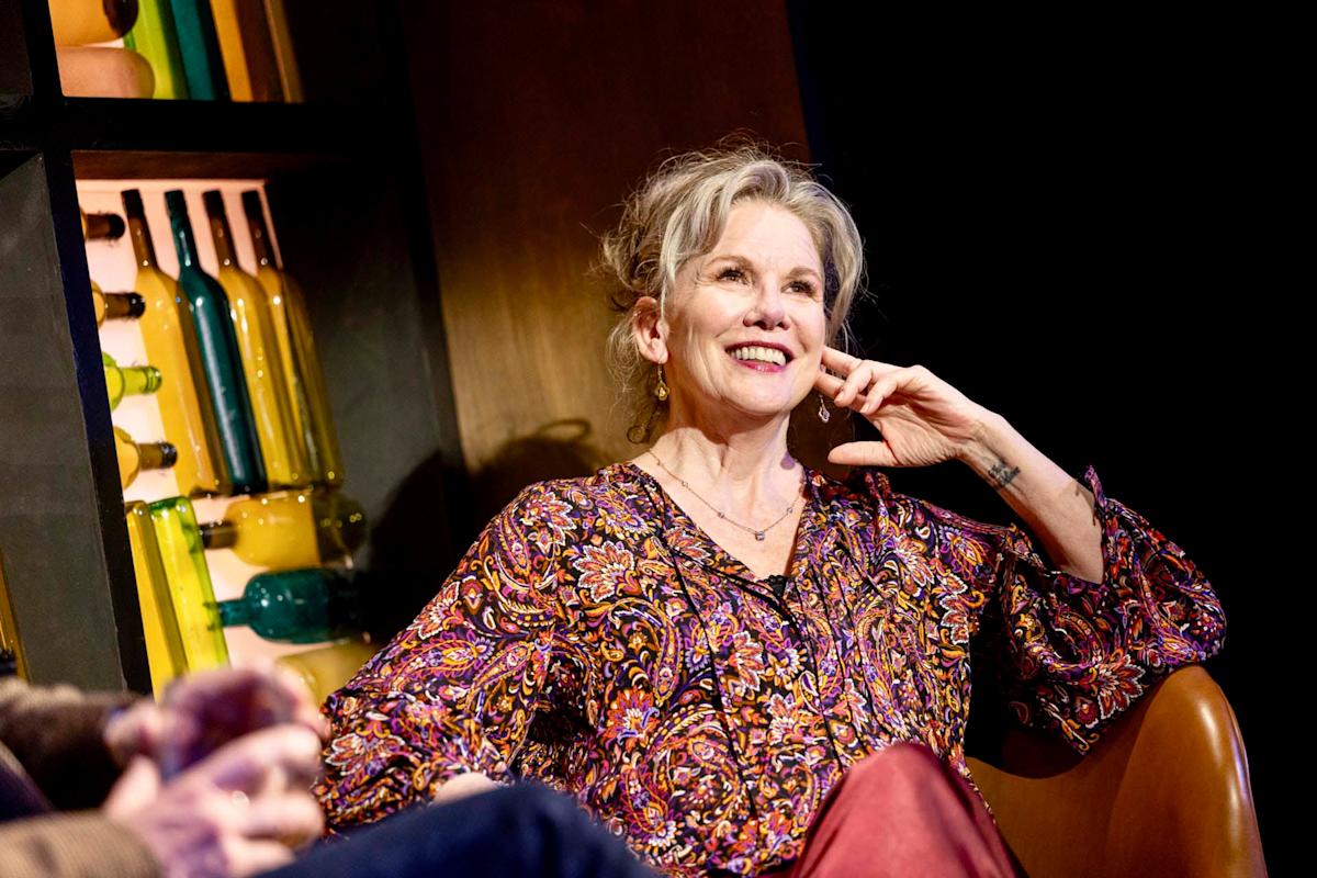 An older woman with gray hair and a patterned blouse smiles while seated in a leather chair. She rests her chin on her hand, with colorful bottles displayed on a shelf behind her.