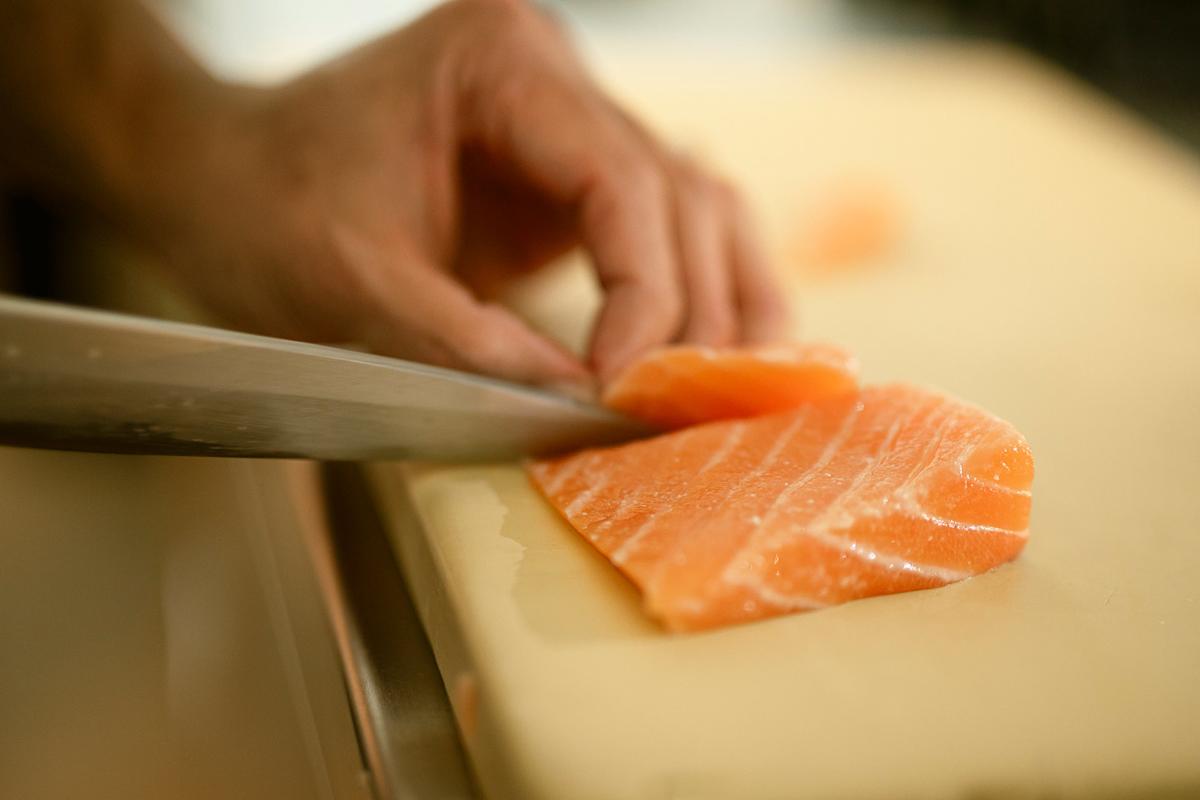 A close-up of a hand slicing a piece of raw salmon with a sharp knife on a light-colored cutting board.