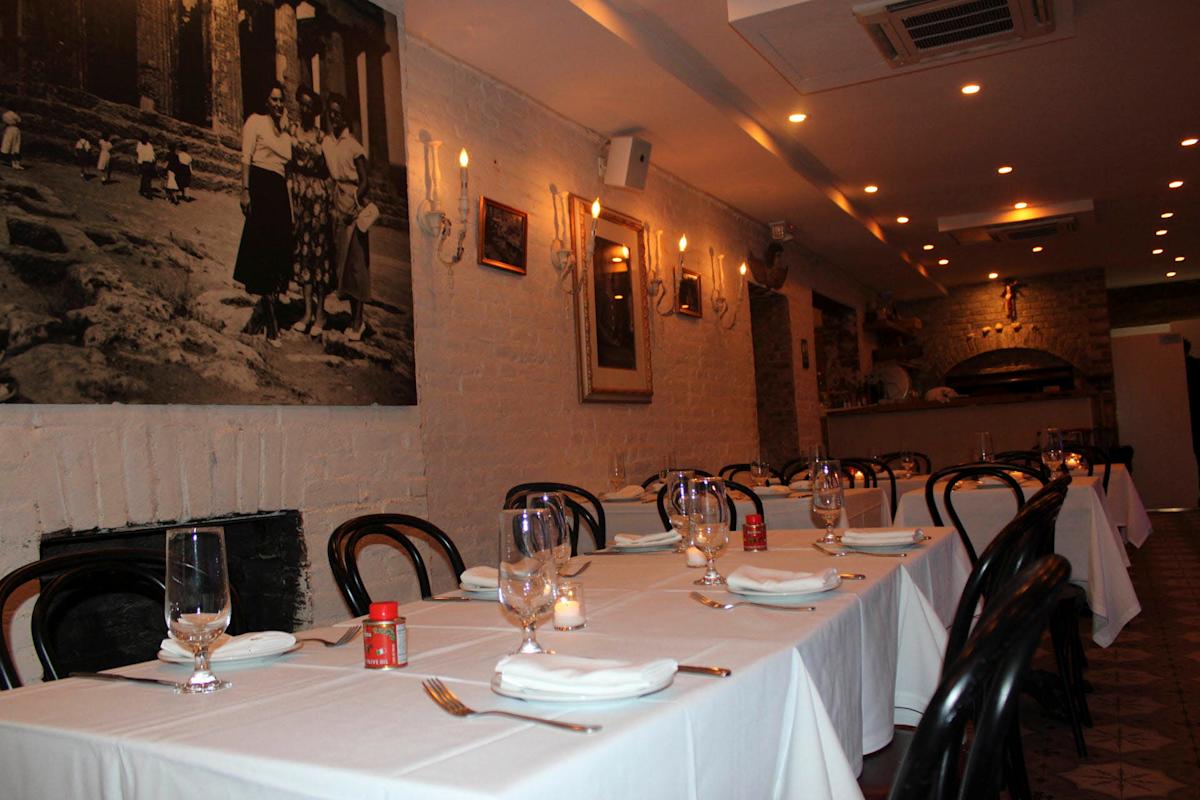 A cozy restaurant interior with neatly set tables draped in white tablecloths, chairs around each table, and a dimly lit ambiance. A large black-and-white photograph hangs on a white brick wall. Candles and glasses are arranged on the tables.