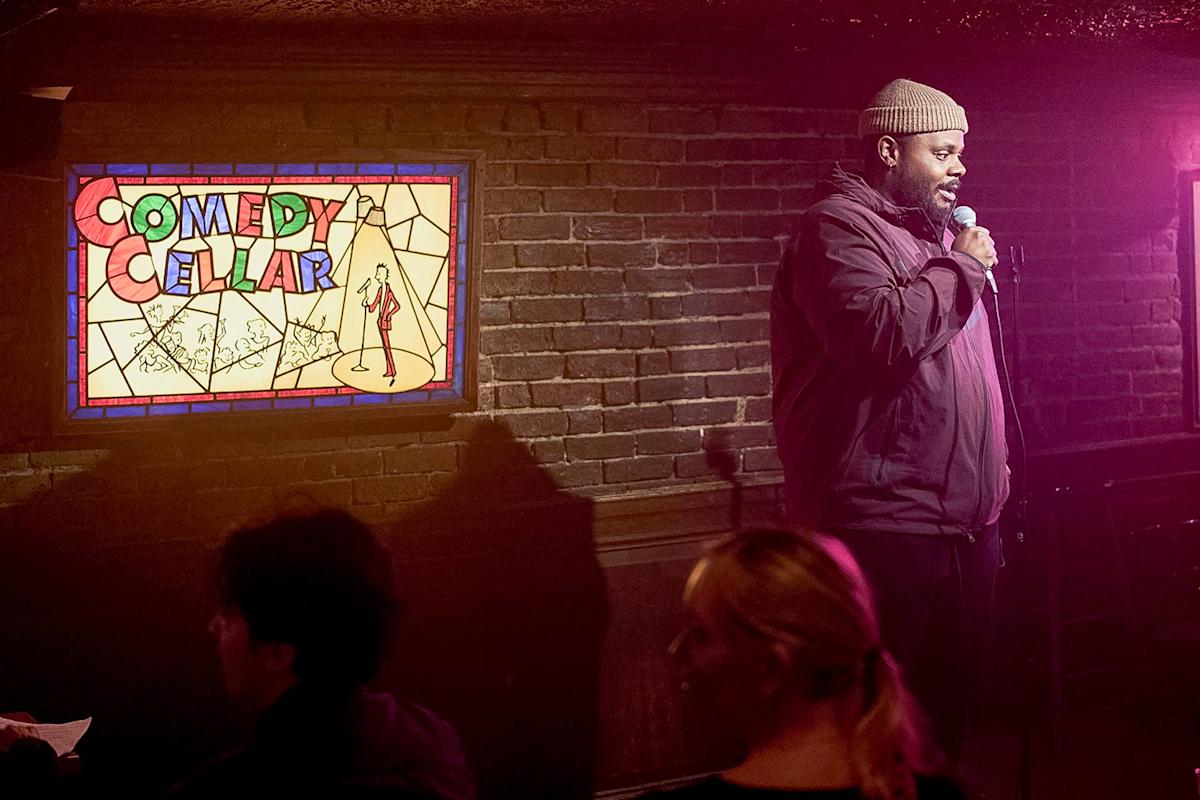 A comedian performs onstage with a microphone at the Comedy Cellar, standing beside a colorful stained-glass sign, while audience members sit and watch in a dimly lit, brick-walled room.