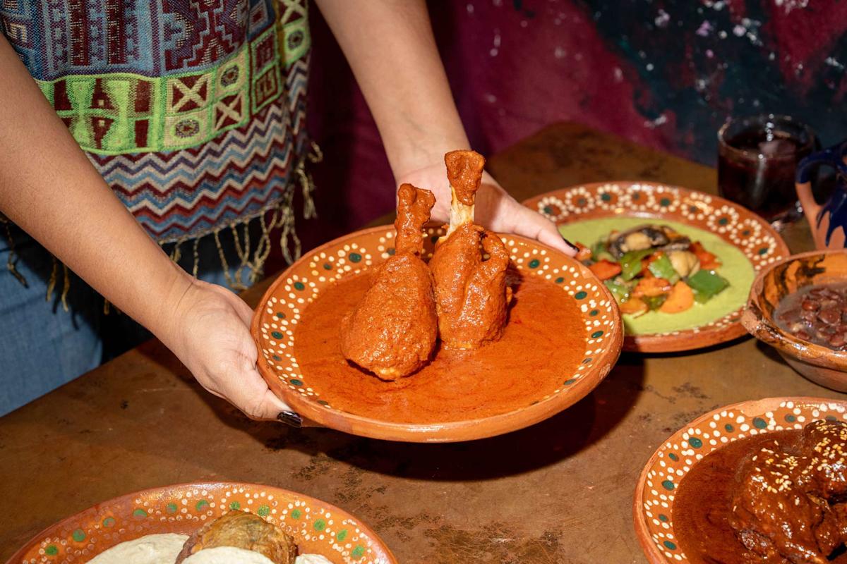 Saavedra holds a traditional clay plate with two chicken drumsticks covered in red mole sauce. Other colorful Mexican dishes are on the wooden table.