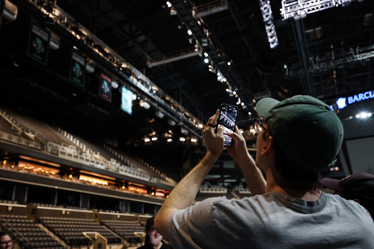 A person wearing a green cap takes a photo or video with a smartphone inside a large, mostly empty sports arena with bright overhead lights and tiered seating.