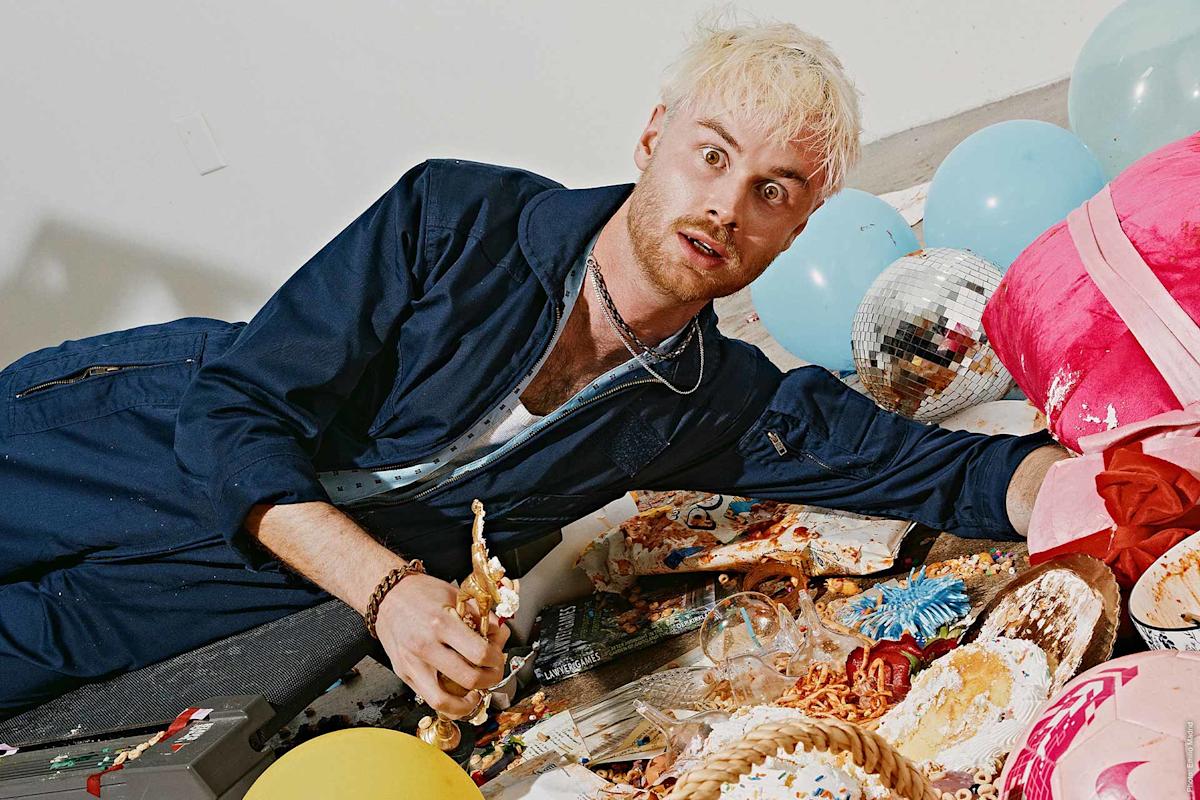A man with bleached blond hair and a blue jumpsuit lies on the floor among party balloons, a disco ball, and a mess of food and decorations, holding a small gold trophy and looking surprised.
