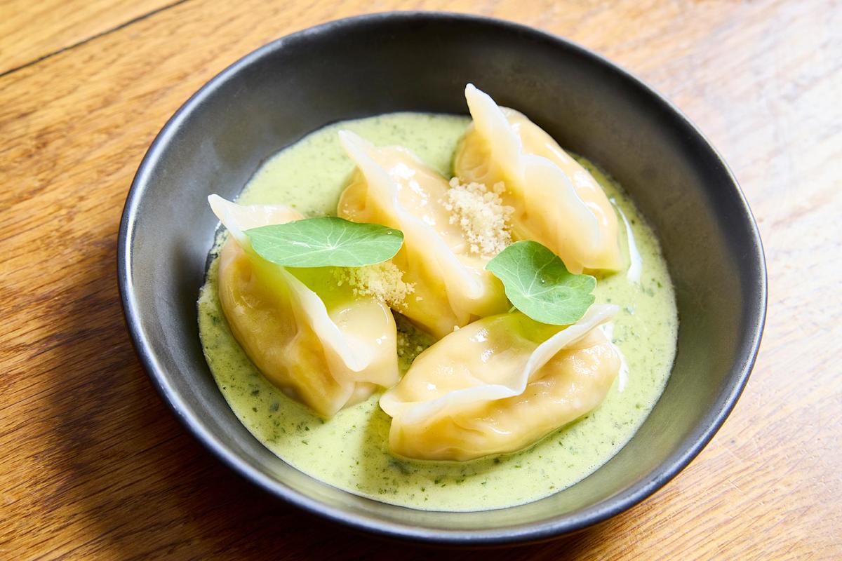 Four dumplings served in a black bowl, resting in a creamy green sauce, garnished with grated cheese and two fresh green leaves, on a wooden surface.