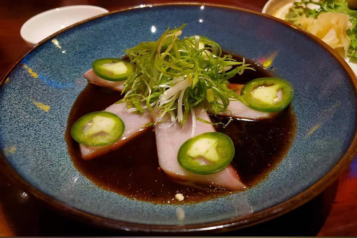 Sashimi slices topped with jalapeño and shredded greens are arranged in a blue bowl filled with dark soy-based sauce. A small dish with pickled ginger is partially visible in the background.