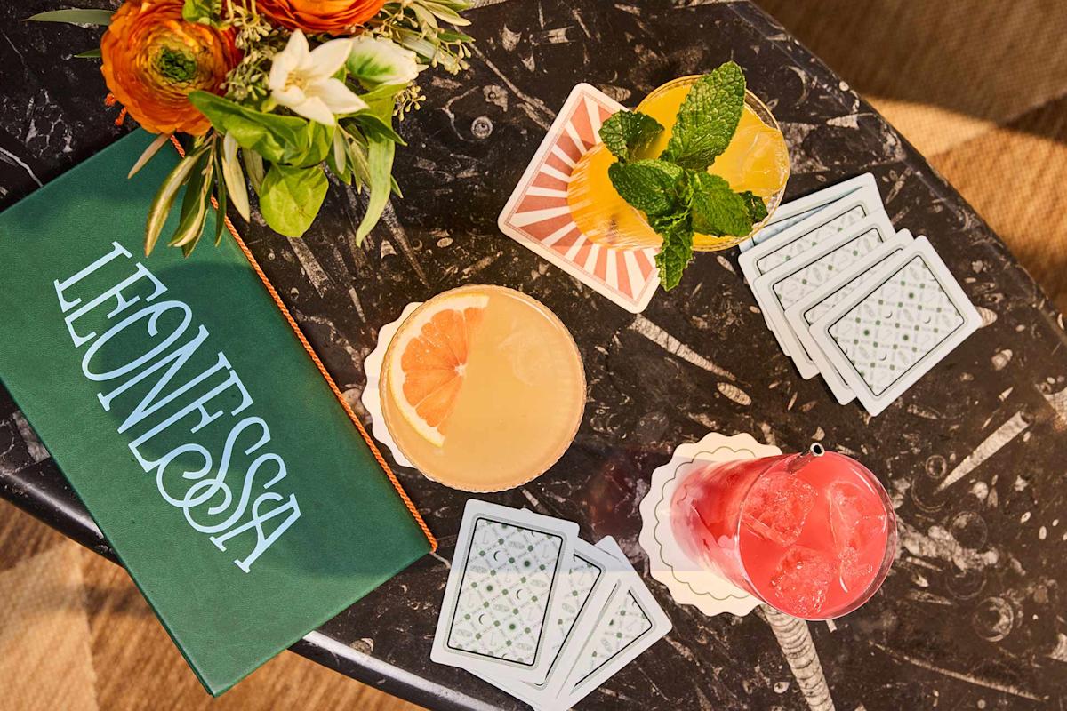 A marble table with three colorful cocktails, a stack of patterned coasters, a bouquet of flowers, and a green menu with "LEONESSA" written on it in white letters.
