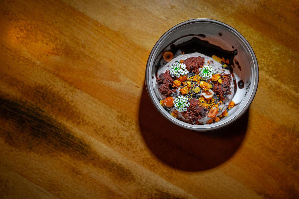A ceramic bowl filled with a chocolate dessert garnished with nuts, edible flowers, and green herbs, placed on a wooden surface with warm lighting casting a shadow.