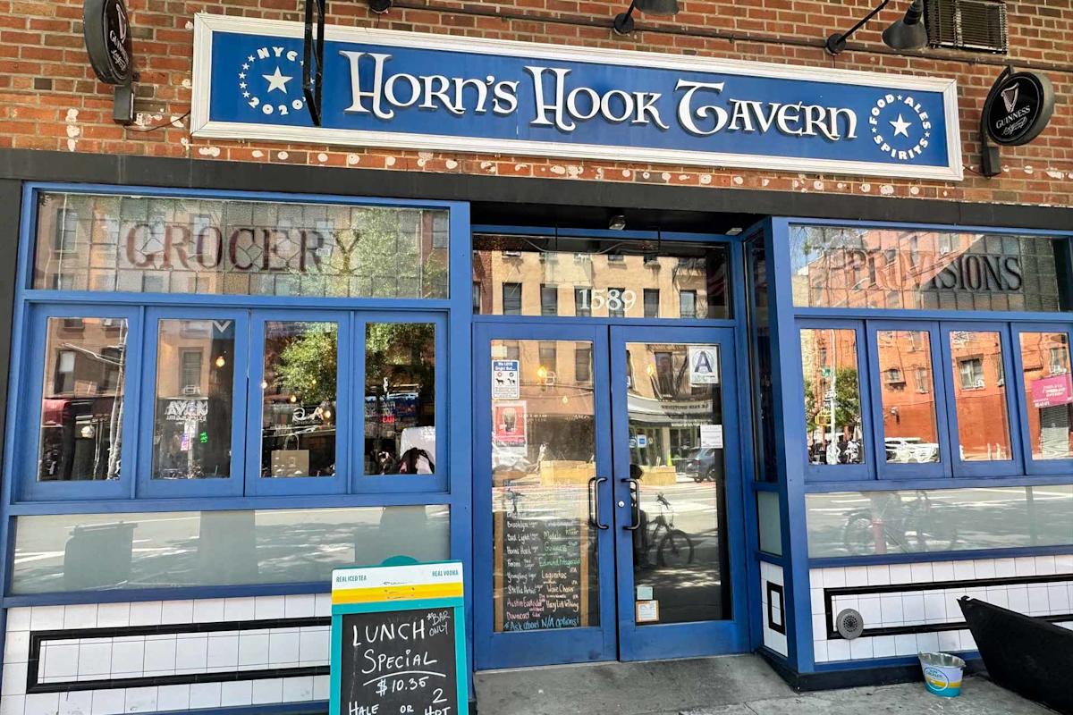Exterior of Horn's Hook Tavern, a blue-fronted bar and restaurant with large windows, a sidewalk chalkboard sign advertising lunch specials, and reflections of buildings across the street.