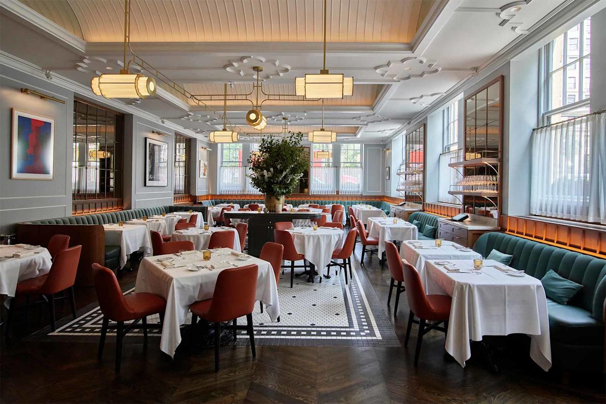 Elegant restaurant interior with white tablecloths, red chairs, and green banquette seating. Large central floral arrangement, modern lighting fixtures, wall art, and tall windows fill the space with natural light.