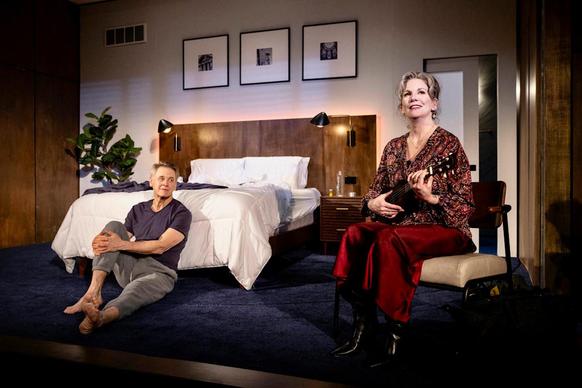 A woman seated on a chair in a bedroom holds a musical instrument, smiling. A man sits on the floor nearby, resting his hands on his knees. The room has a neatly made bed, wooden walls, framed pictures, and a plant.