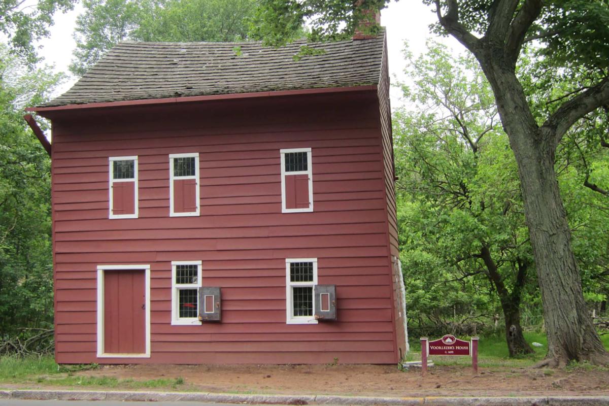A two-story red wooden house with white trim stands by a road, surrounded by green trees. A small sign in front reads “Wallace House & Old Dutch Parsonage.” A logo with a bear is in the top left corner.