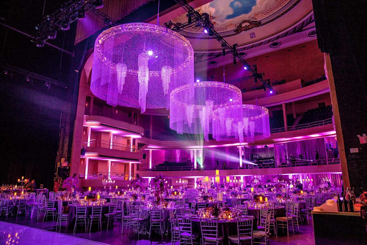 A grand ballroom set for an event, featuring round tables with elegant centerpieces and silver chairs, illuminated by purple lighting and large, shimmering chandelier decorations hanging from the ceiling.