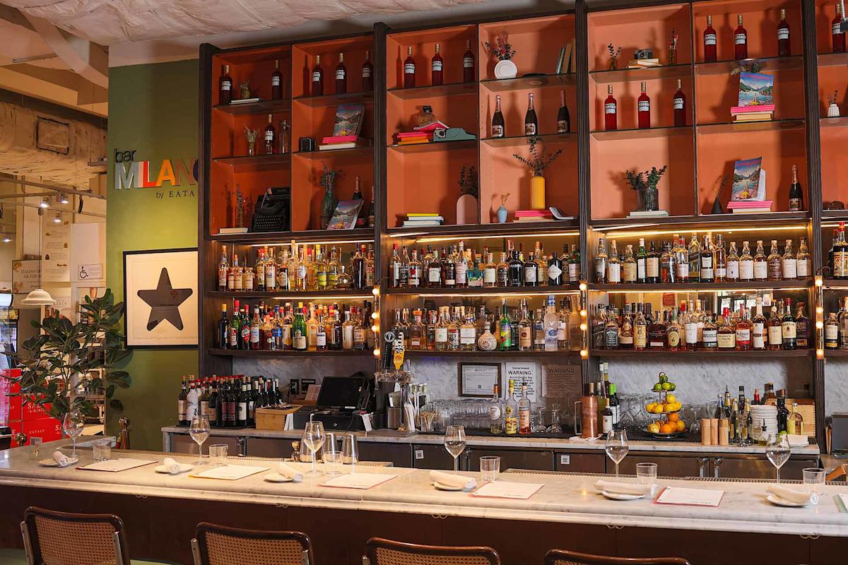 A stylish bar with shelves filled with bottles of liquor and wine, glasses hanging above, a marble counter set with plates and wine glasses, and chairs lined up in front. Warm lighting creates a welcoming atmosphere.