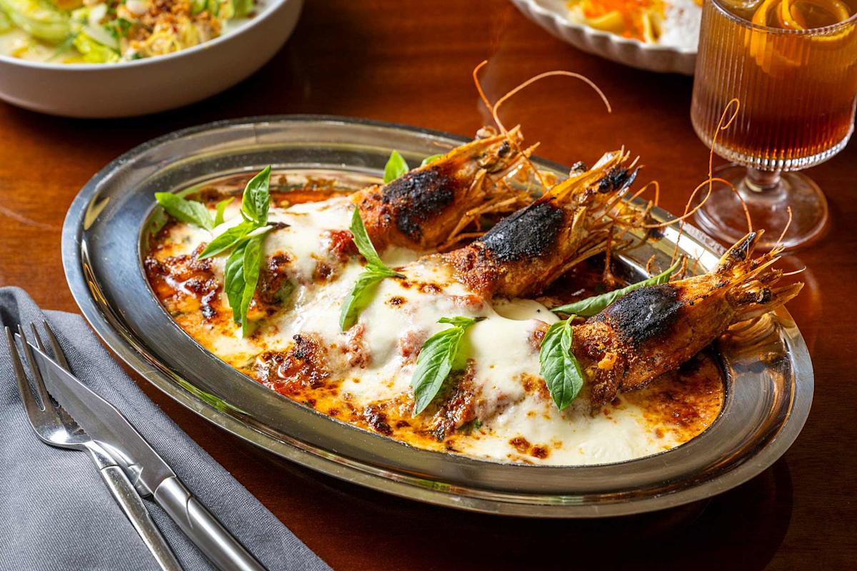 A silver platter with baked prawns in melted cheese and tomato sauce, garnished with fresh basil leaves. A glass of iced drink and a salad are in the background on a wooden table.