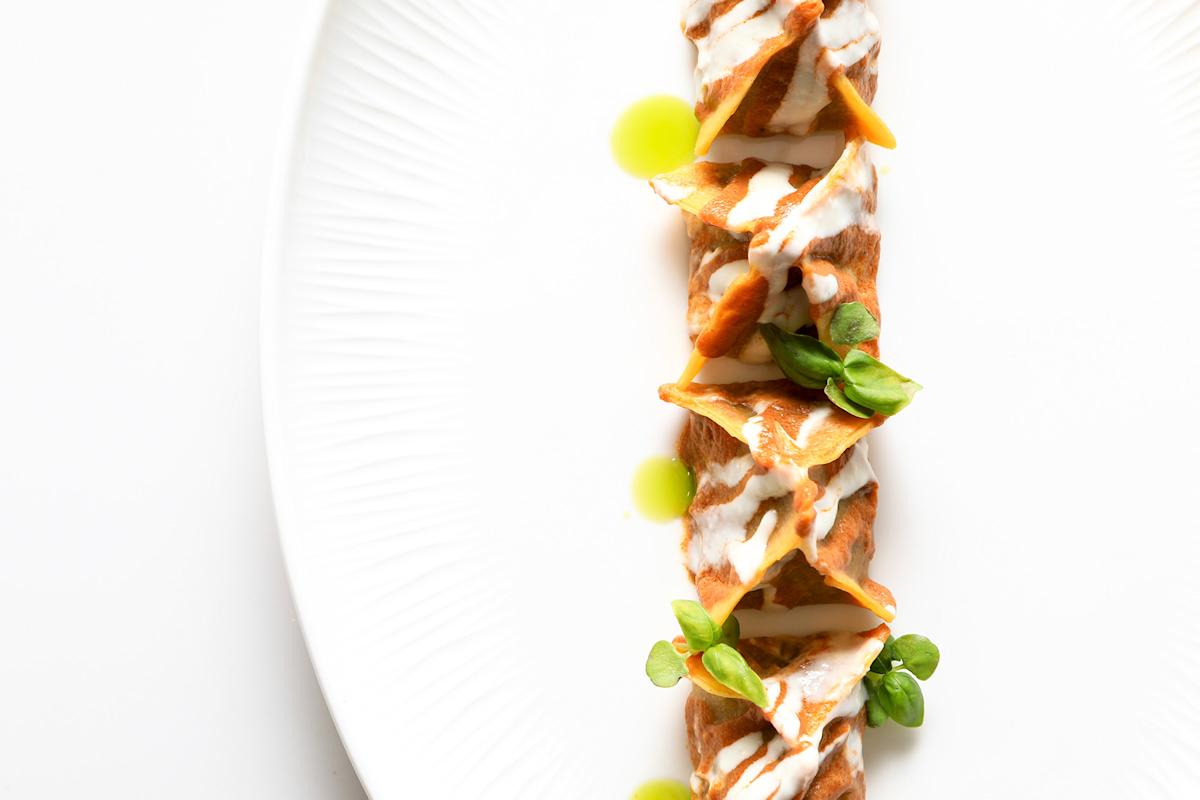 Four neatly arranged dumplings topped with a creamy white sauce and green herbs are presented on a white plate, garnished with small drops of green oil for a minimalist, elegant look.