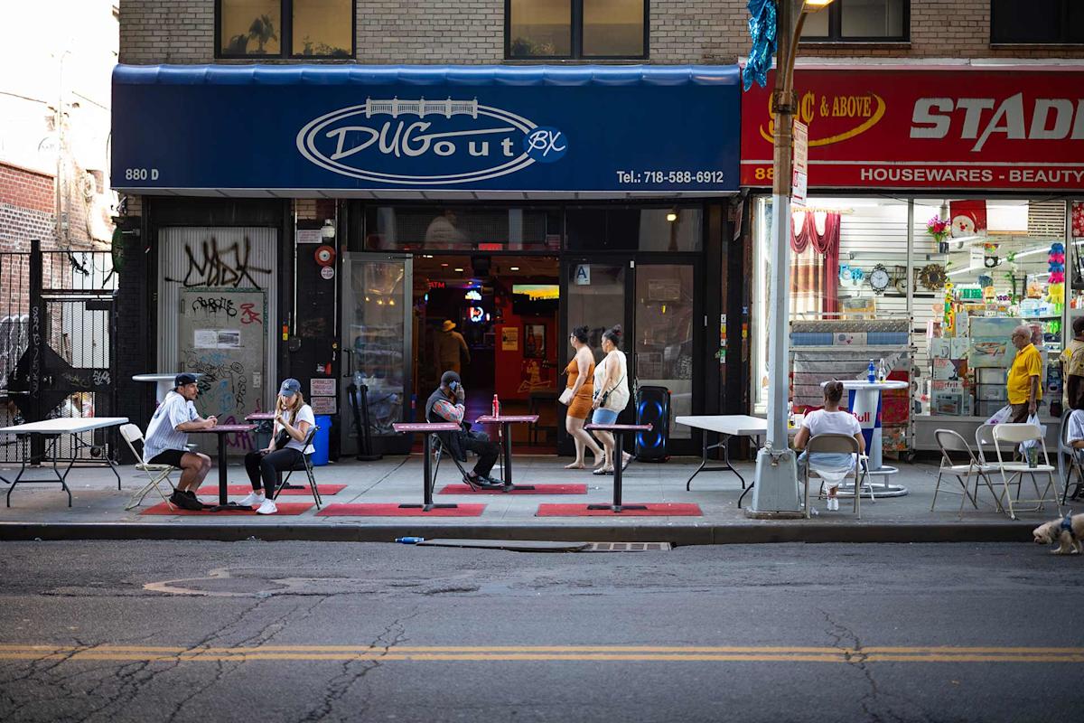 People sit at tables on a sidewalk outside Dugout bar; others walk past. The bar has a blue awning, and a brightly lit store selling housewares is next door. The street is quiet with little traffic.