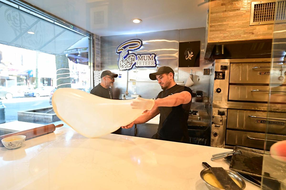 Two people work in a pizzeria kitchen; one is stretching pizza dough by hand on a counter, while the other observes. Large ovens and a neon "Mum Feter" sign are visible in the background.