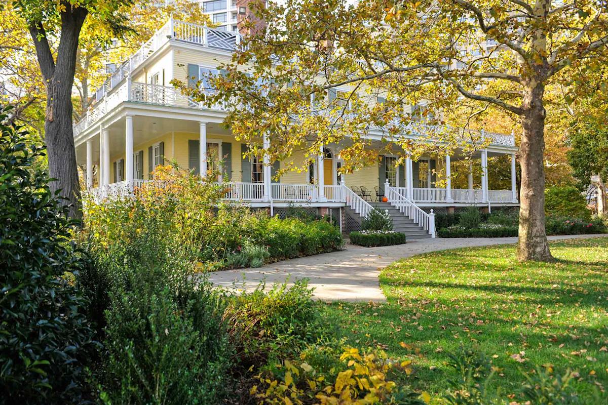A large yellow house with a wraparound porch is surrounded by trees and lush greenery. A curving pathway leads to the front steps. The scene is bathed in sunlight, highlighting the vibrant colors of the foliage.