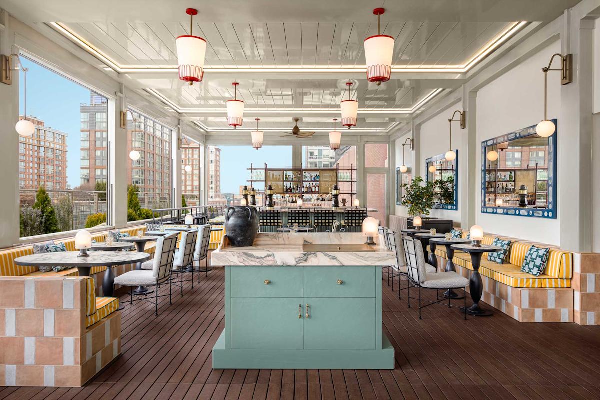 An elegant rooftop restaurant with marble tables, striped yellow booth seating, modern white chairs, a central island, and a bar at the back, surrounded by large windows with city views.