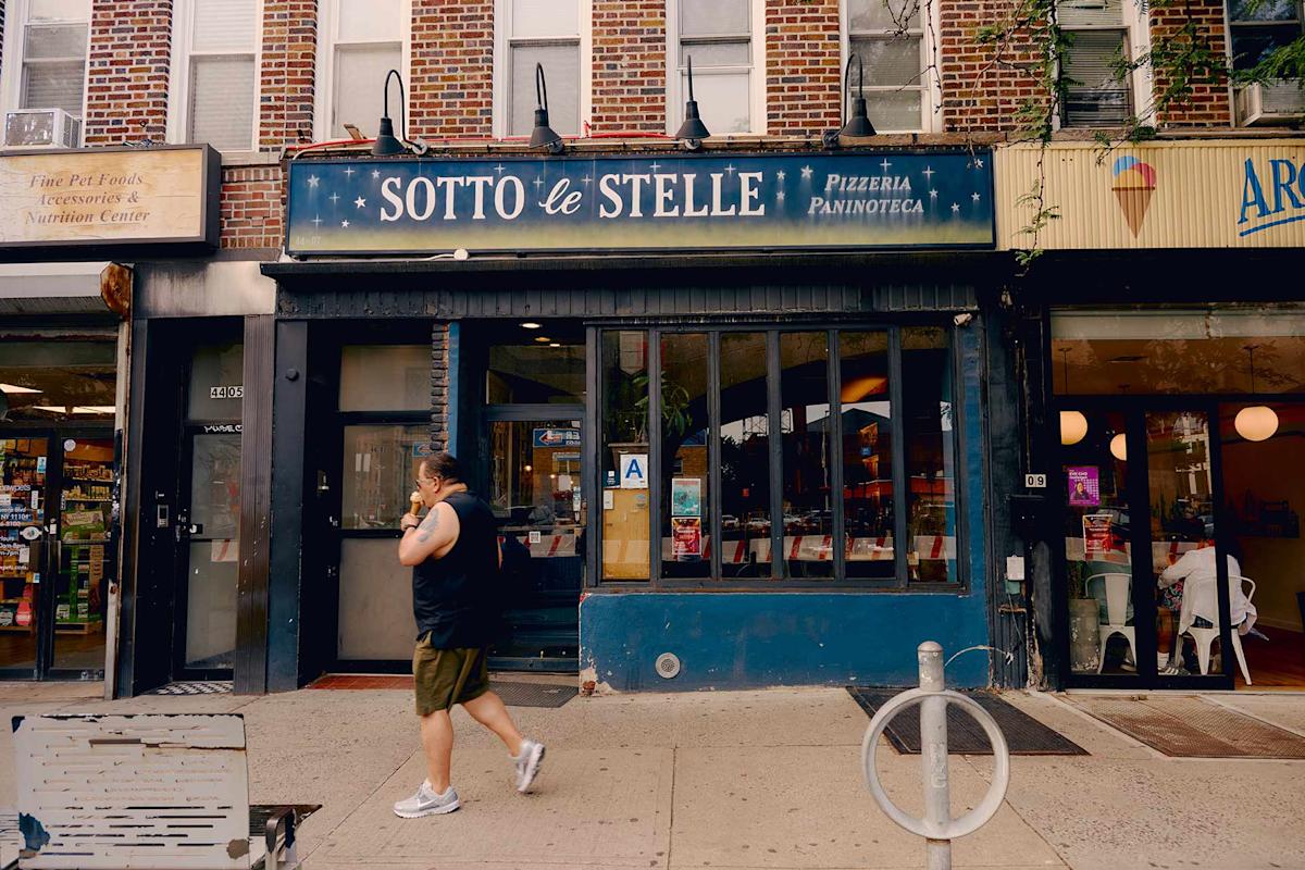A person walks past the storefront of “Sotto le Stelle,” a pizzeria and paninoteca on a city street, with brick buildings and other shops visible on either side.