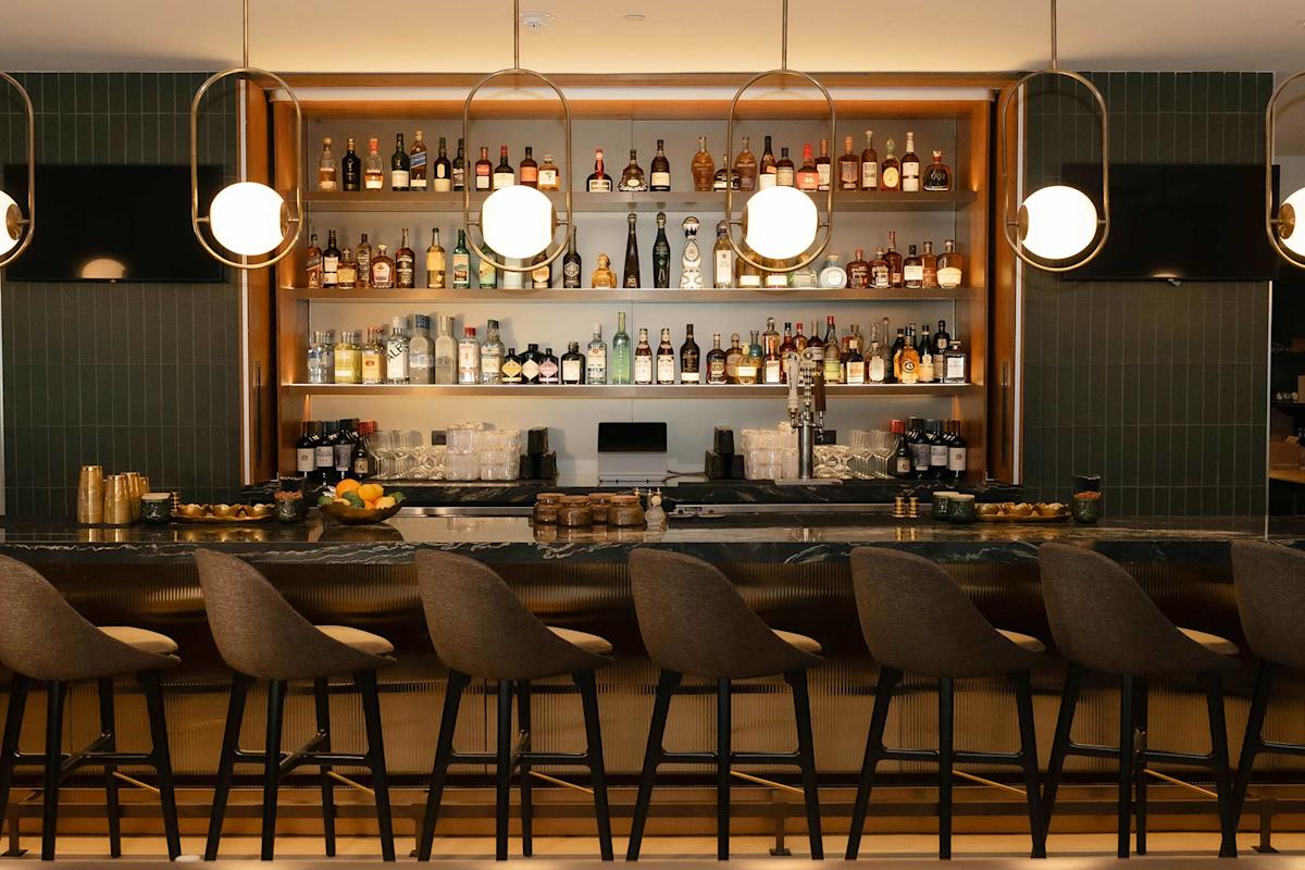 A modern bar with high-backed chairs, globe pendant lights, and shelves stocked with assorted liquor bottles behind a long, marble countertop.