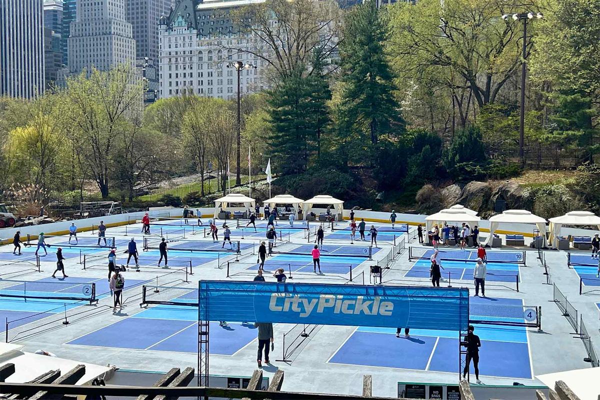Multiple people play pickleball on several blue outdoor courts surrounded by trees in a city park, with tall buildings visible in the background. A banner reads "CityPickle" in the foreground.