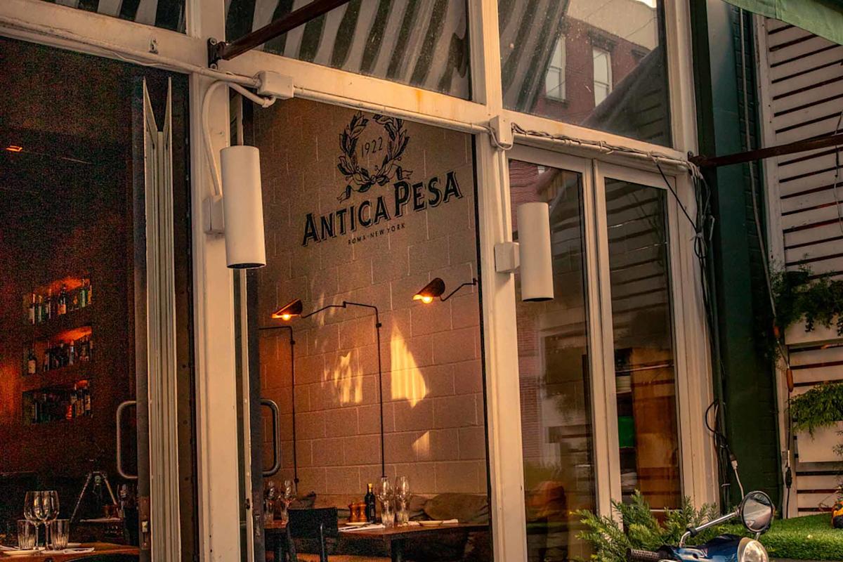 The image shows the exterior of Antica Pesa restaurant, with glass windows revealing a warmly lit interior, tables set for dining, and the restaurant’s name and logo visible on the inside wall.